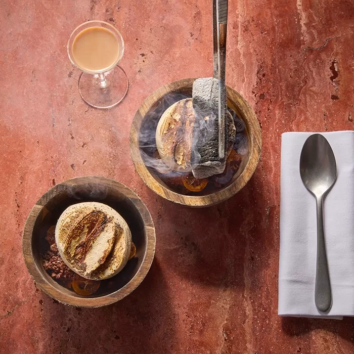 Two wooden bowls with scoops of ice cream and sauce on a reddish marble surface. One bowl has a smoking hot stone held by tongs above it. A drink in a glass and a spoon on a white napkin are nearby.