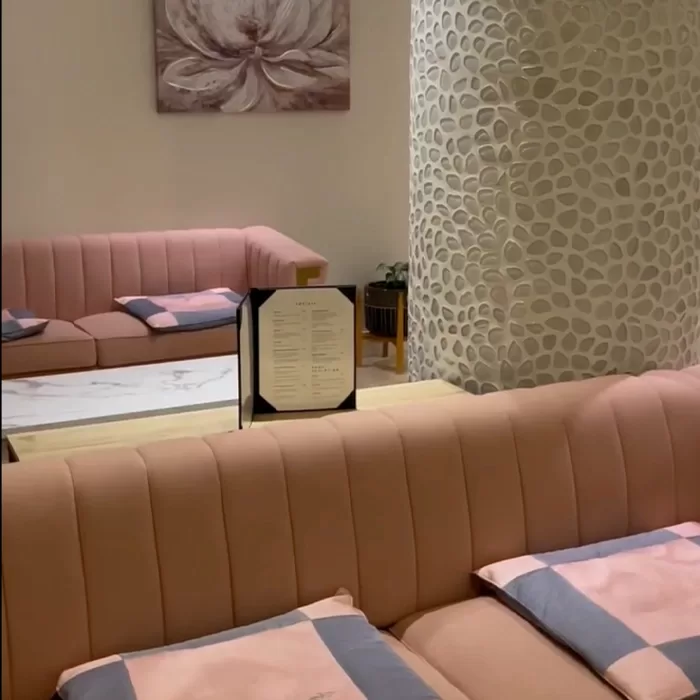 A cozy lounge area with pink cushioned sofas, pink and gray pillows, a marble coffee table with a menu standing upright, a potted plant, a large floral wall art, and a decorative pillar with a leaf pattern.