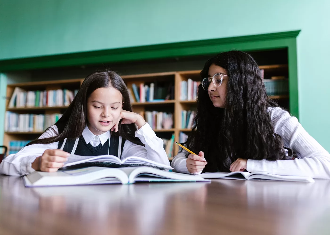 Two girls sit at a desk in a library, reading and studying together with open books in front of them. One girl is reading while the other looks on, holding a pencil. Shelves of books are visible in the background.