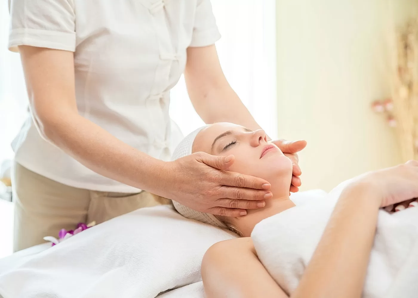 A person receiving a facial massage while lying on a spa bed. Another person in a white uniform gently massages their face. The setting is calm and serene, emphasizing relaxation and wellness.