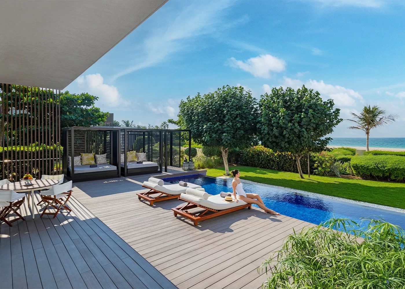 A woman relaxes on a lounge chair by an outdoor pool overlooking the ocean. The scene includes lush greenery, modern cabanas, and a small dining table on a wooden deck under a clear blue sky.