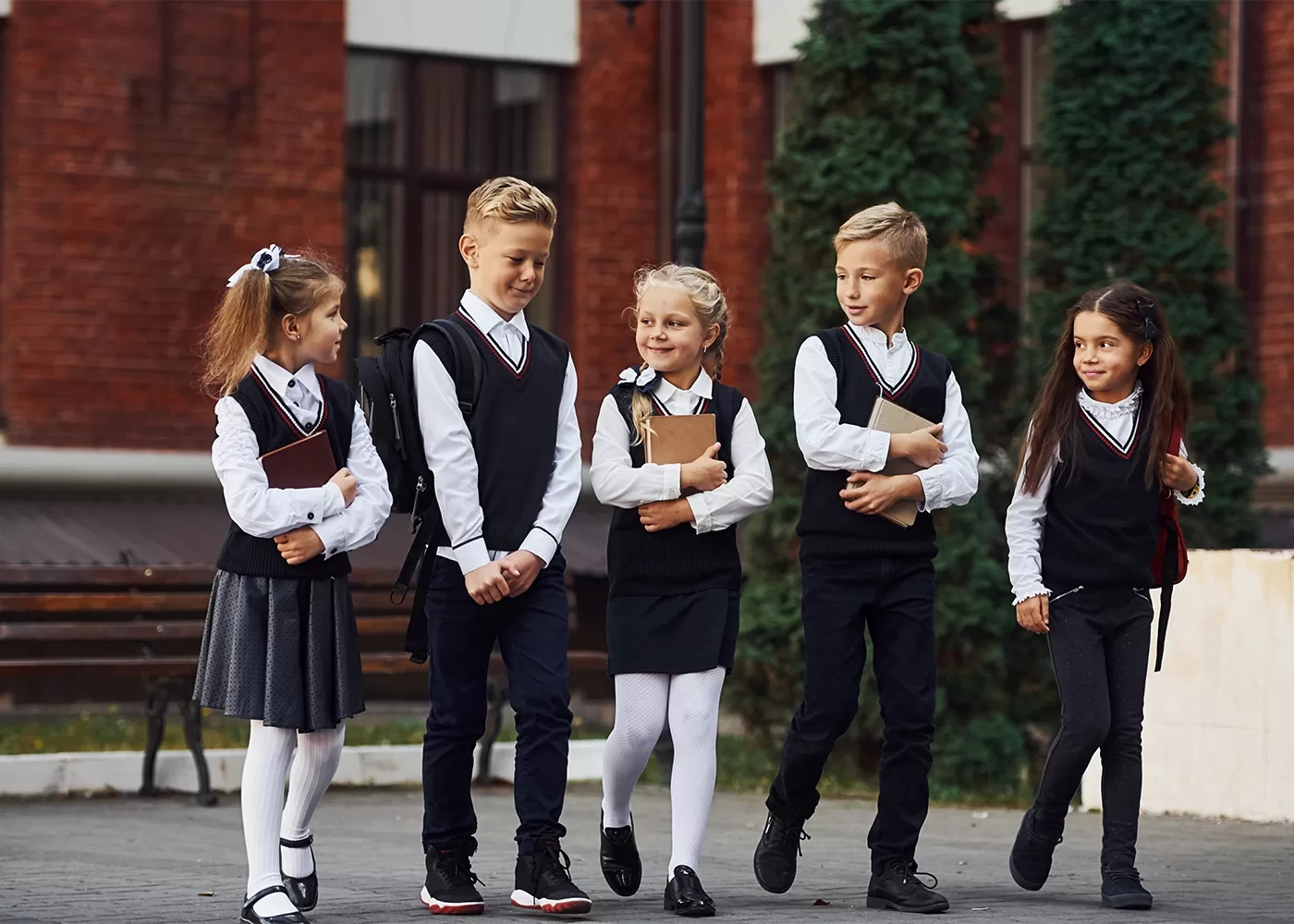 Five young children in school uniforms walk together outside, talking and smiling. They carry books and backpacks, with a red-brick building and green trees in the background.