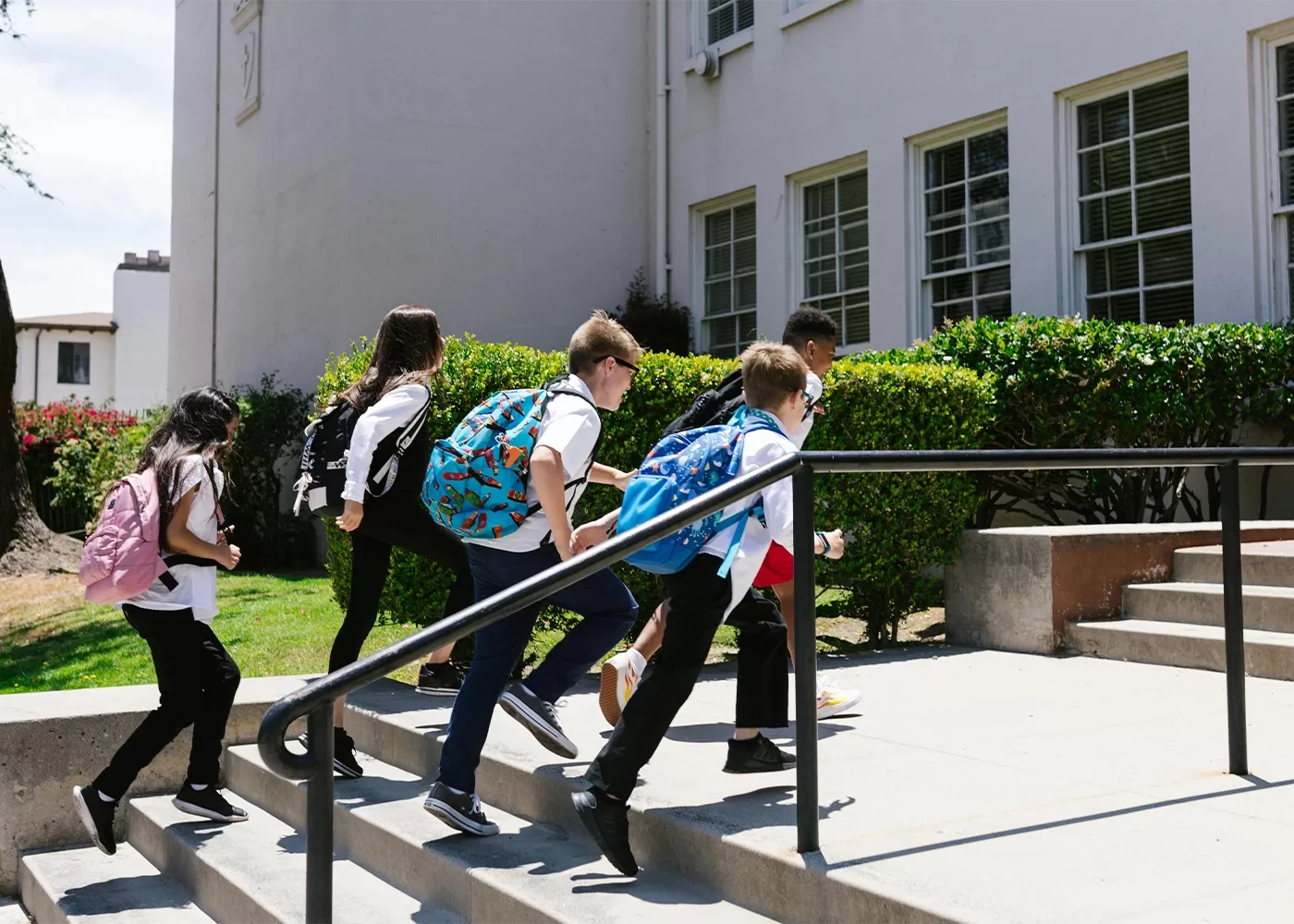 Five children with backpacks walk up the steps toward a school building with large windows and green bushes outside, appearing to head inside together.