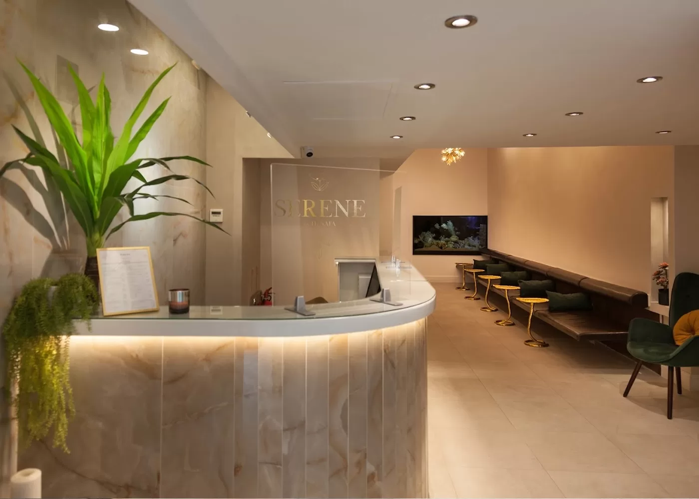 A modern spa reception area with a curved marble desk, accented by a large plant and a small picture frame. Behind, a hallway with plush green seating and golden stools leads to a softly lit waiting area, featuring a large fish tank.