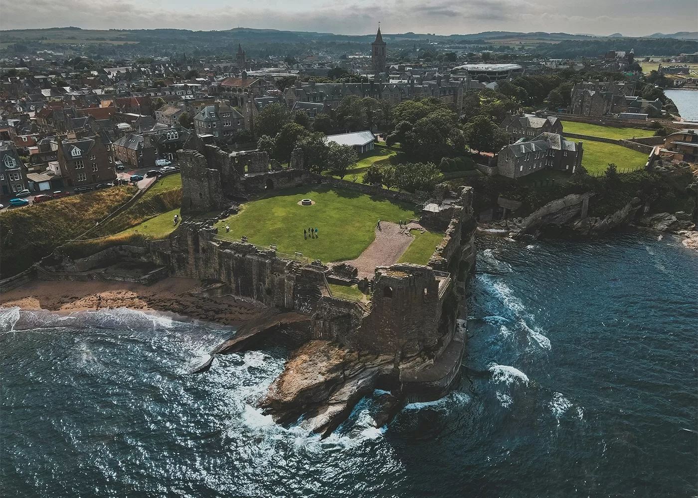 Aerial view of coastal ruins of a large stone castle, surrounded by green lawns, beside the sea. A historic town with many stone buildings stretches out behind the castle under a cloudy sky.