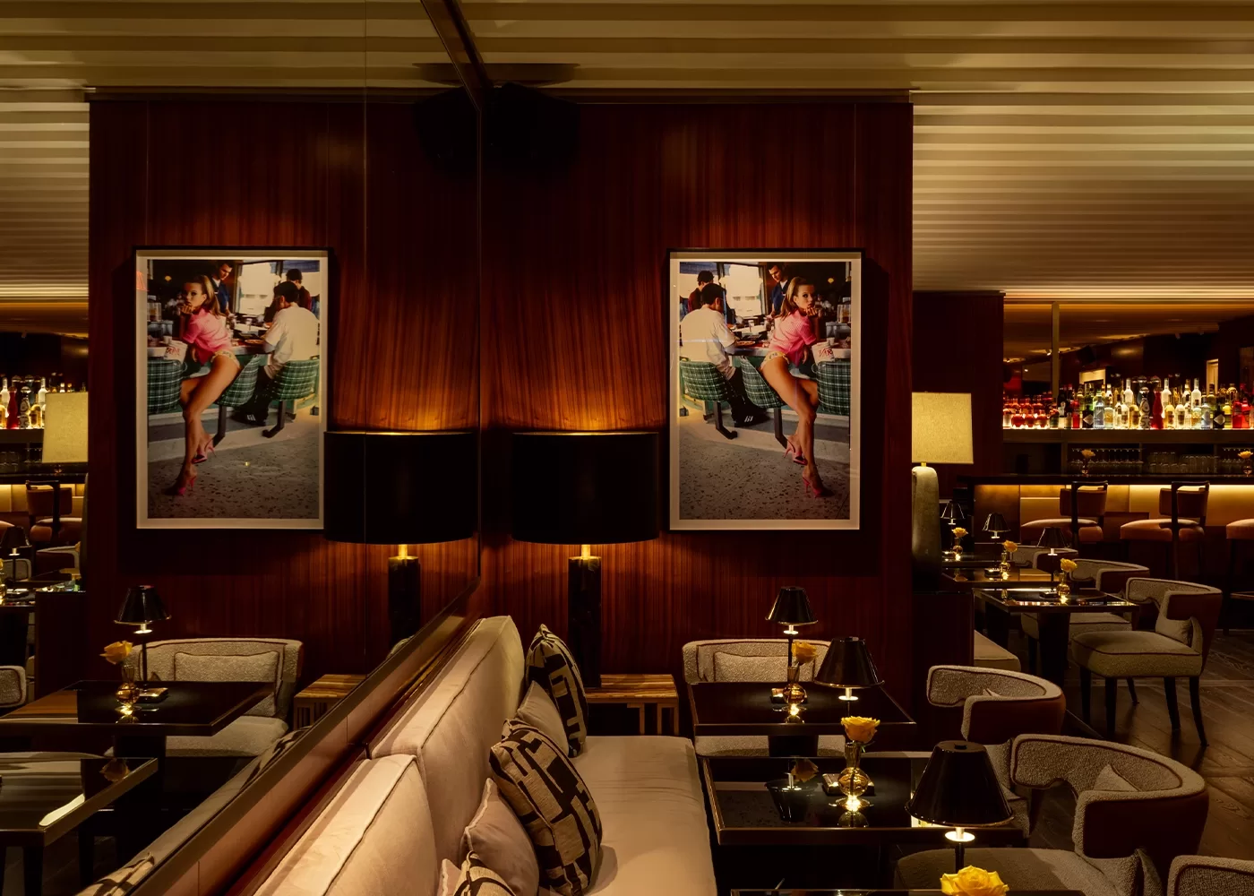 A dimly lit, elegant lounge with plush seating and polished wood paneling. The walls are adorned with large, framed photographs, and several small black tables are set with lamps and glassware, creating an inviting, sophisticated ambiance.