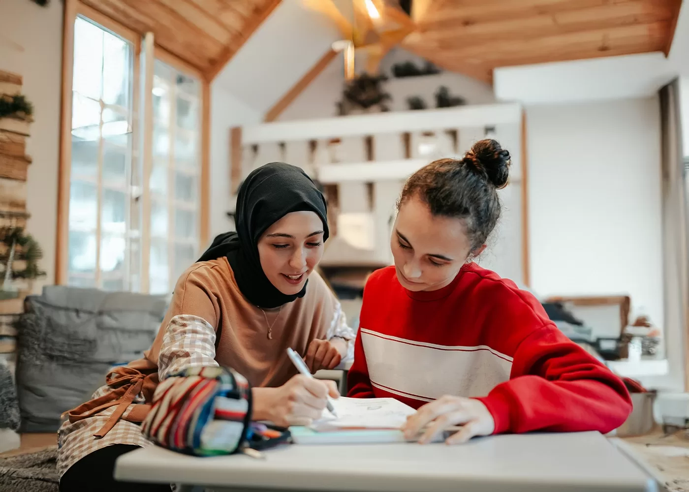 Two young women sit at a table in a cozy room. One wears a hijab and writes in a notebook, while the other, in a red sweater, observes. They appear focused and engaged in a study session.