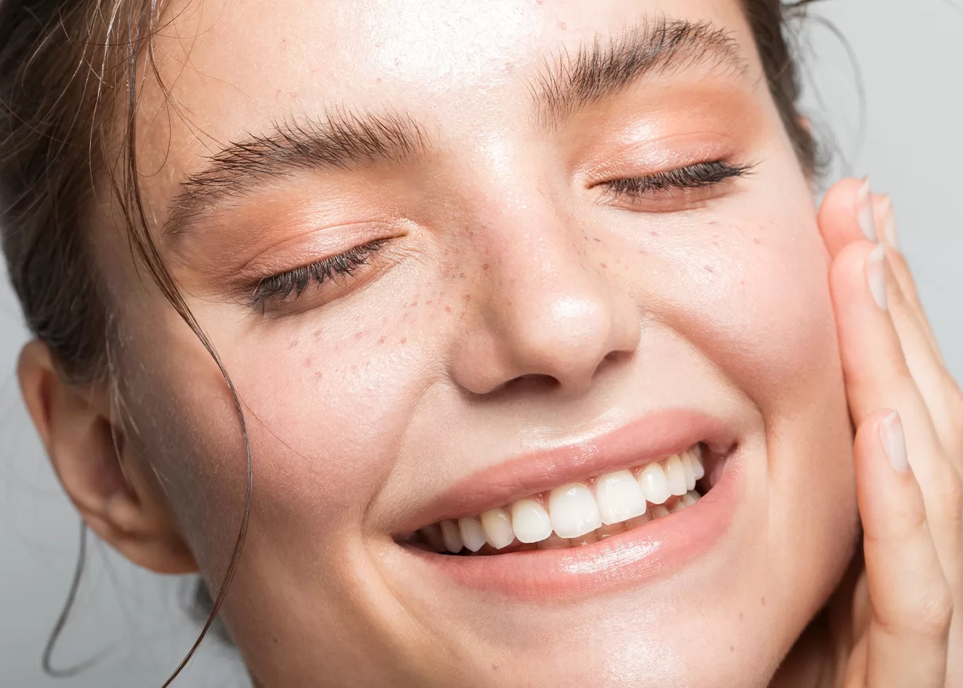 A close-up of a person smiling with closed eyes. They have clear skin with light freckles and are wearing peach-toned eyeshadow. Their hand gently touches their cheek. Their expression conveys a sense of joy and relaxation.