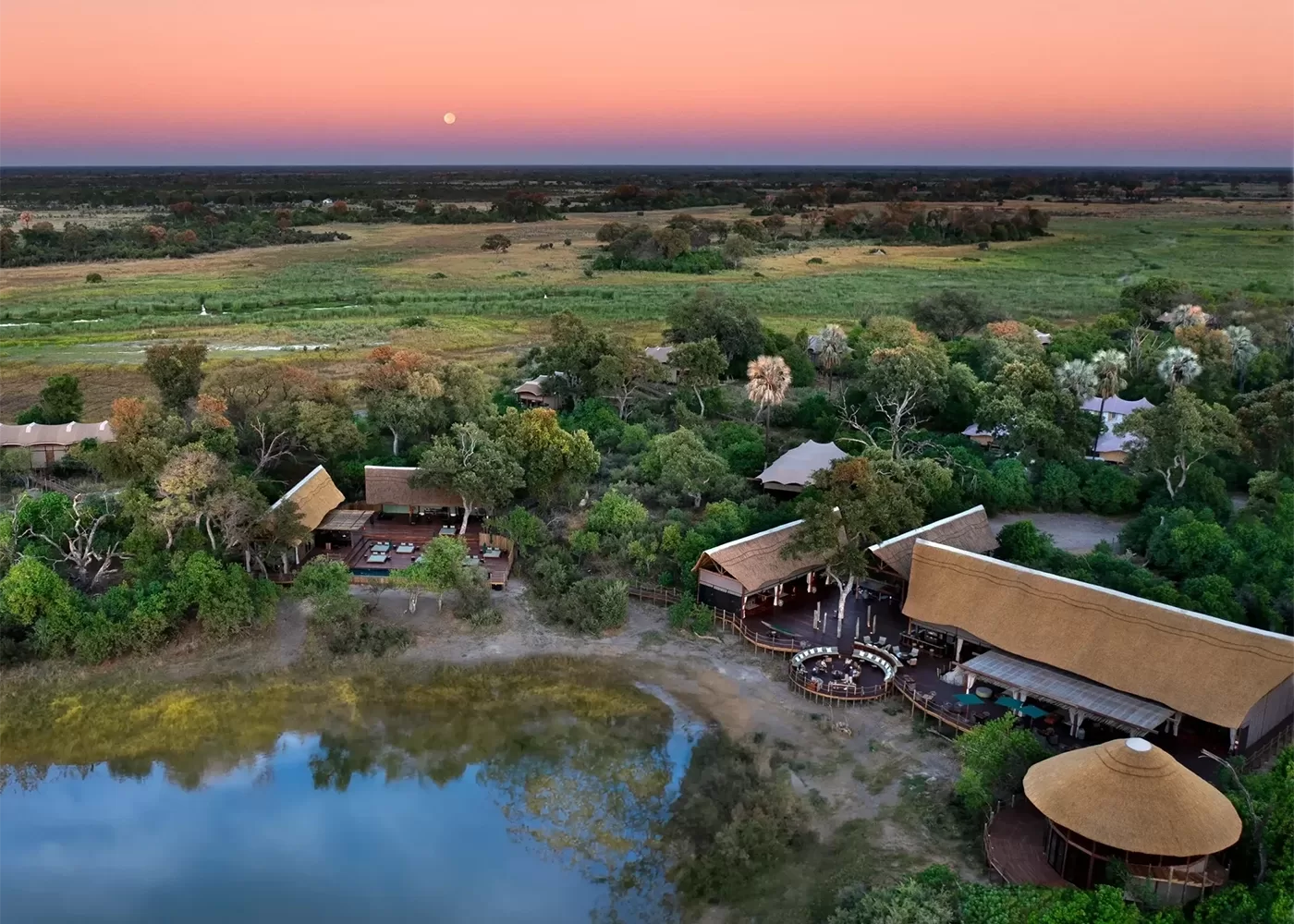 Aerial view of a safari lodge with thatched roofs surrounded by lush greenery and a calm lagoon. Open plains stretch into the distance under a pink and orange sunset sky. A circular outdoor seating area is visible.