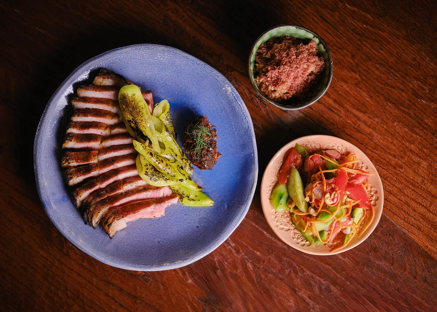 Aerial view of a wooden table with a blue plate Blythburg chop, pork fat, and smoked chilli relish served at AngloThai in London.