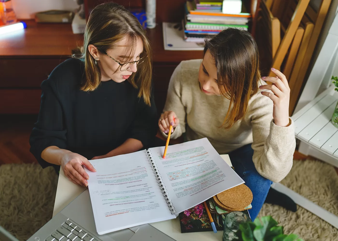 Two people are sitting at a table, focused on studying. They are looking at a notebook filled with highlighted text. One person is pointing at the notes with a pencil, and the other is listening attentively. Books and stationery are scattered around.