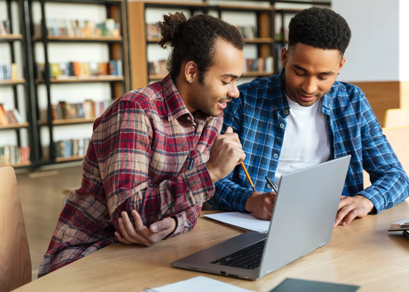 Two people sitting at a library table, working on a laptop. One is wearing a red plaid shirt, holding a pencil, and looking at the screen, while the other in a blue plaid shirt writes in a notebook. Shelves of books are visible in the background.