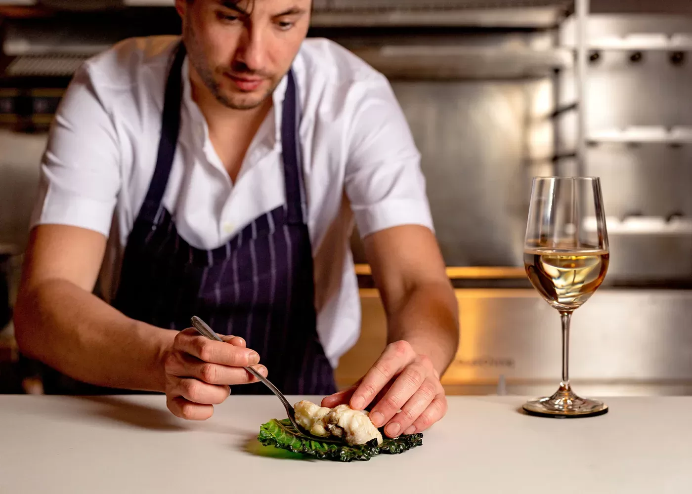 A chef in a white shirt and striped apron carefully plates a dish in a kitchen. A glass of white wine is placed on the counter beside the dish, illuminated under warm lighting at Kebab Queen, Soho, London.