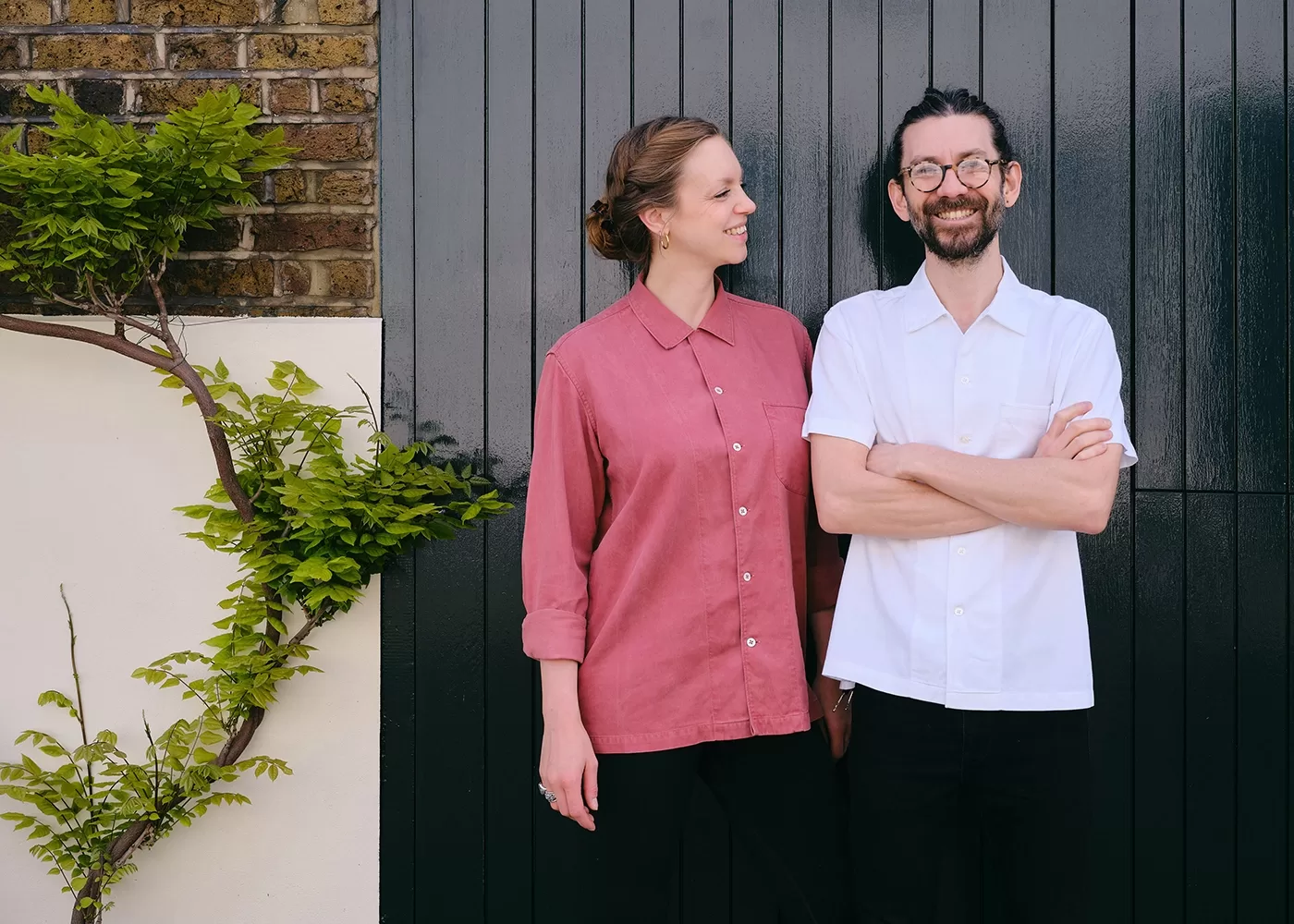 A woman in a red shirt smiles and looks at a man in a white shirt who stands with arms crossed. They are in front of a dark vertical wooden panel, with a green leafy plant climbing the wall beside them - the people are Desiree and John Chantarasak, chef's of the new restaurant AngloThai in London.