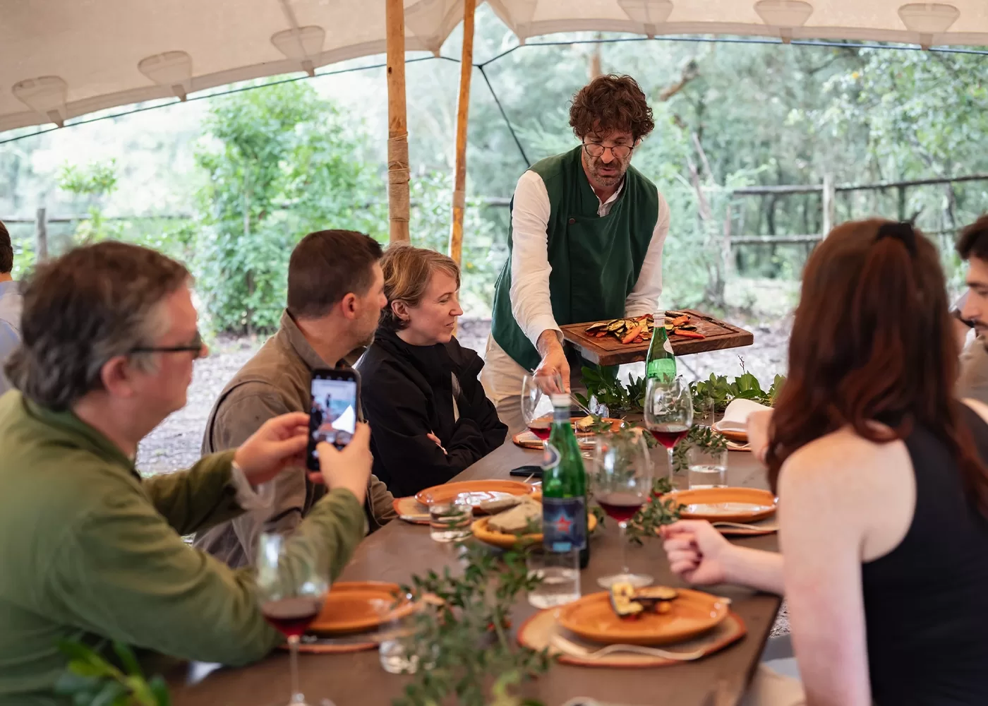 A group of people sit around a wooden table under a canopy, enjoying a meal outdoors. A server presents a platter of food. Wine glasses, dishes, and foliage decorate the table. One person is taking a photo with a smartphone.