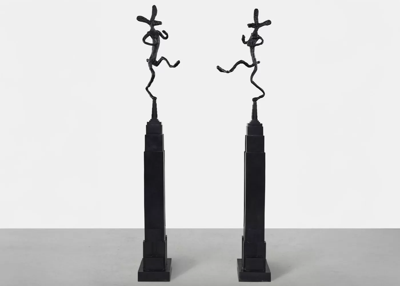 Two tall, slender black sculptures stand side by side, each topped with a whimsical, abstract figure in a dynamic pose. The design is minimalistic and stands on a flat surface with a plain background - Empire State with Bowler, Mirrored, 1997 by Barry Flanagan.