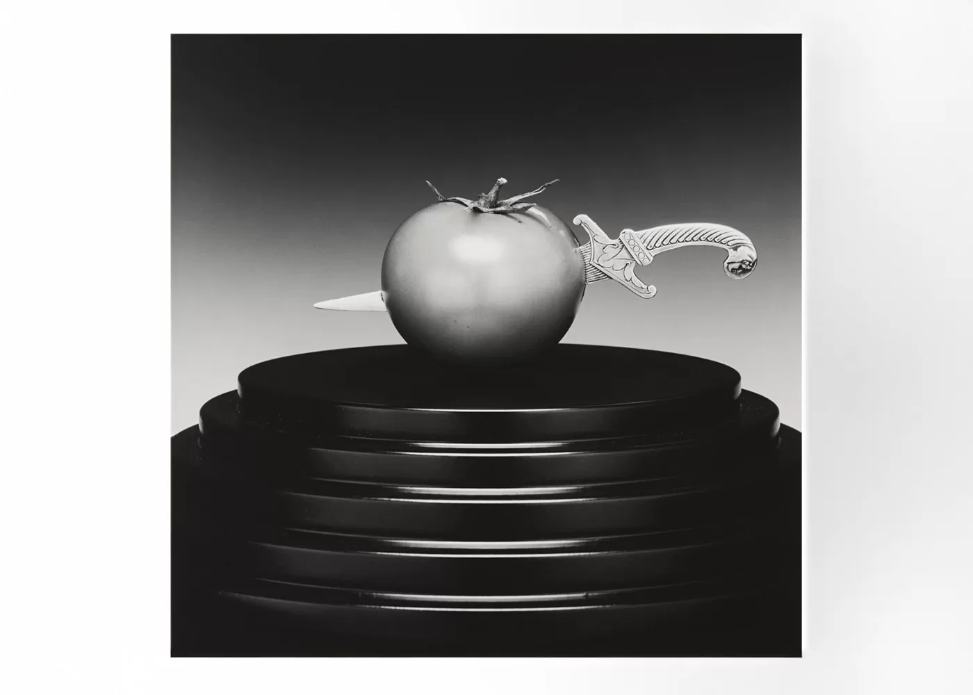 A black and white photograph of a tomato skewered by a decorative knife, displayed on a multi-tiered pedestal. The image contrasts the smooth tomato and intricate knife design against the stark background - Tomato/Knife, 1989 by Robert Mapplethorpe.