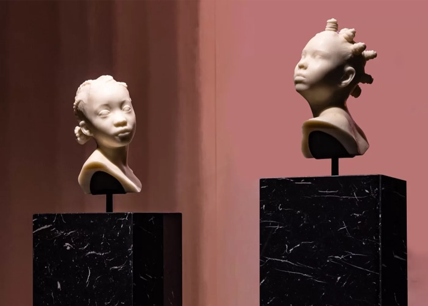 Two sculpted busts on black marble pedestals, depicting stylized portraits of children with unique hairstyles. The background features a warm, muted gradient - Ebony In Ivory III, 2023 and Ebony In Ivory II, 2023 by Esmaa Mohamoud.