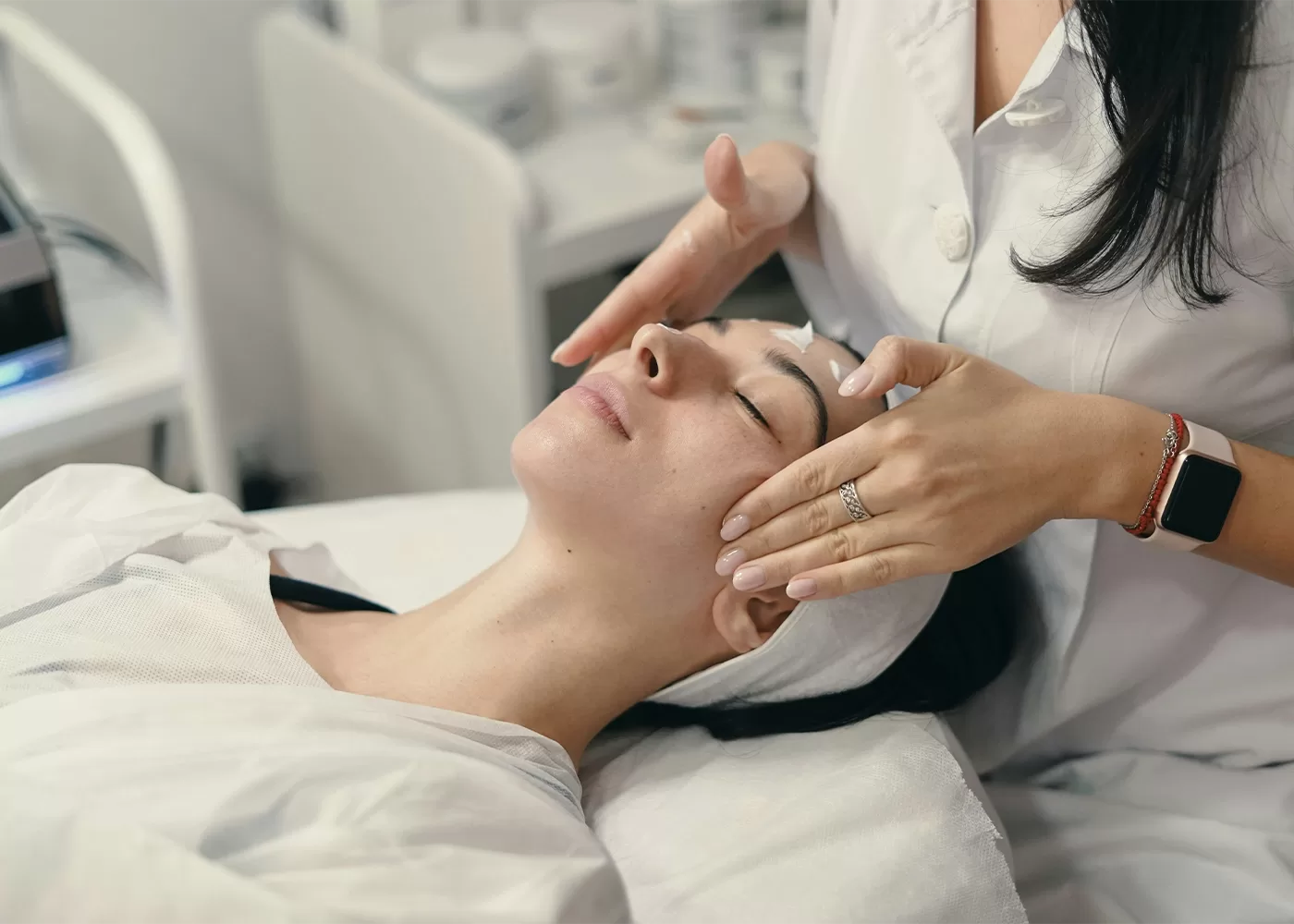 A person receiving a facial treatment in a spa setting. They are lying down with eyes closed, while a professional in a white coat gently massages their temples. Creams and skincare products are visible in the background.