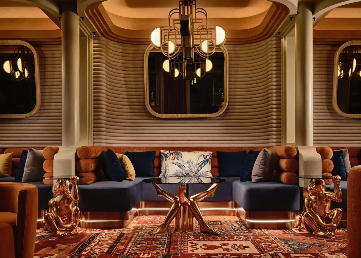 Stylish lounge with a luxurious interior featuring a navy blue sectional sofa, ornate chandelier, geometric wall patterns, a glass coffee table with a star-shaped gold base, and gold dog sculptures. Richly patterned rug covers the floor.