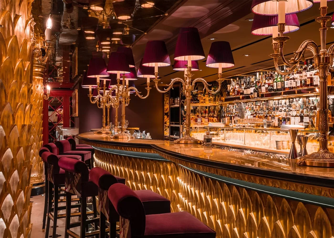 A luxurious bar with gold accents, purple velvet barstools, ornate lamps with purple shades, and a mirrored ceiling. Shelves behind the bar are stocked with various bottles of alcohol.