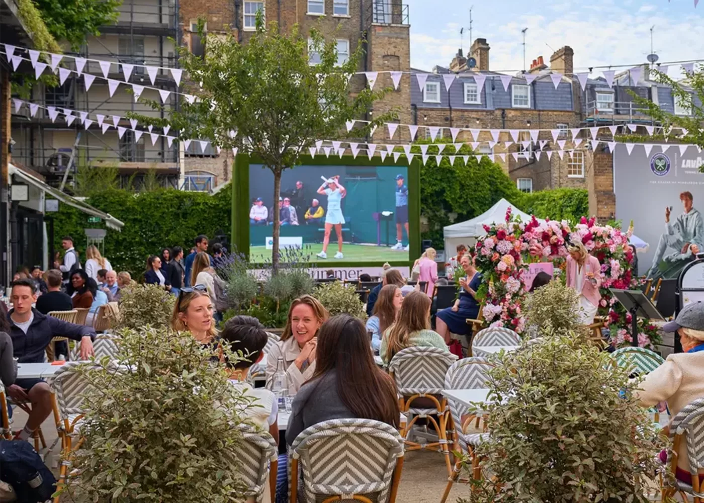 People sit at outdoor tables in a garden setting, watching a large screen showing a tennis match. The area is decorated with bunting and flowers, creating a festive, social atmosphere. Buildings and greenery are in the background.