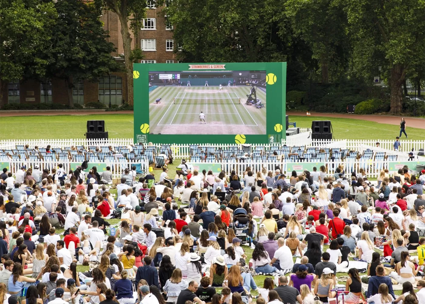 A large crowd sits on grass watching a live tennis match on a giant outdoor screen in a park, surrounded by trees and buildings. The atmosphere is lively and festive.