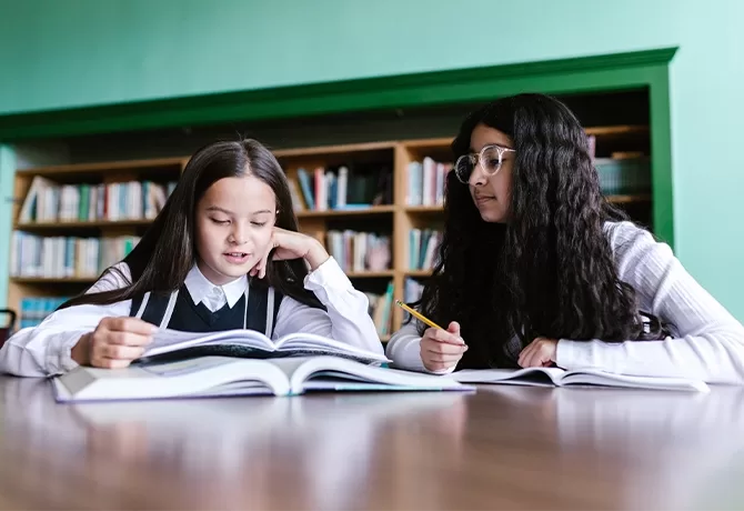 Two girls sit at a table with open books in a library. One girl is reading while the other listens and holds a pencil. Bookshelves filled with books are visible in the background.