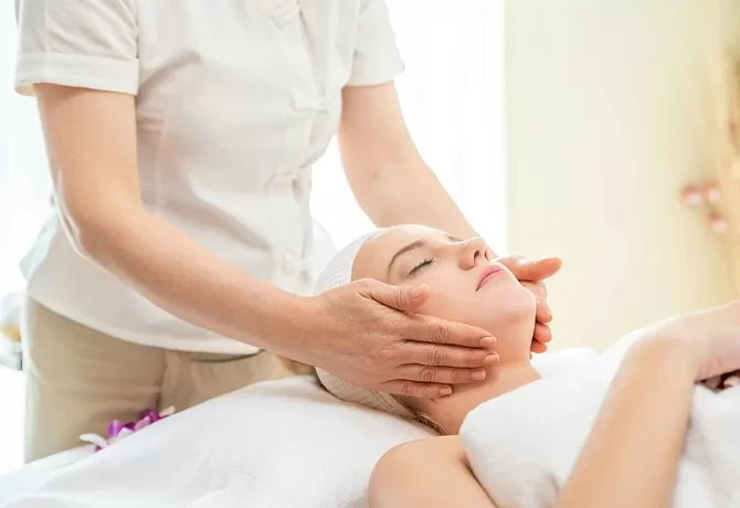 A person receiving a facial massage while lying on a spa bed. Another person in a white uniform gently massages their face. The setting is calm and serene, emphasizing relaxation and wellness.