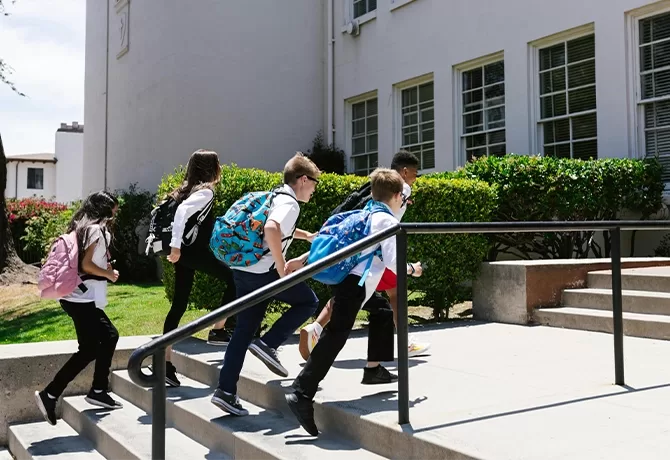 Five children wearing backpacks walk up the steps outside a school building on a sunny day. The school has large windows and is surrounded by green bushes.