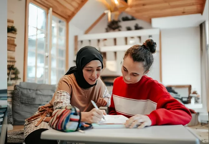 Two young women sit at a table in a cozy room. One wears a hijab and writes in a notebook, while the other, in a red sweater, observes. They appear focused and engaged in a study session.
