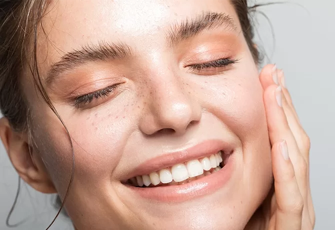 A person with fair skin and freckles smiles with eyes closed, showcasing their radiant complexion. Their makeup features peach-toned eyeshadow and a natural lip color. One hand gently touches their cheek, conveying a sense of contentment and skincare.