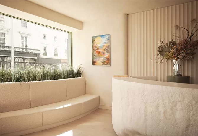 A cozy reception area with a curved beige sofa, a rounded stone desk, and a vase of dried flowers. A large window with reeds offers a view of a street. A colorful abstract painting hangs on the wall, adding a vibrant touch to the neutral decor.