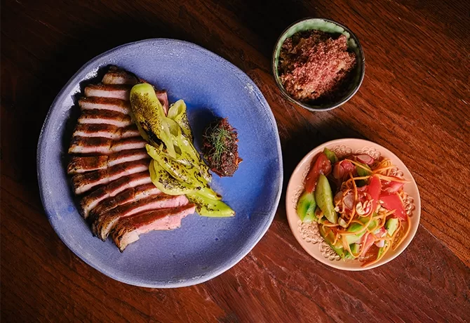 Aerial view of a wooden table with a blue plate Blythburg chop, pork fat, and smoked chilli relish served at AngloThai in London.