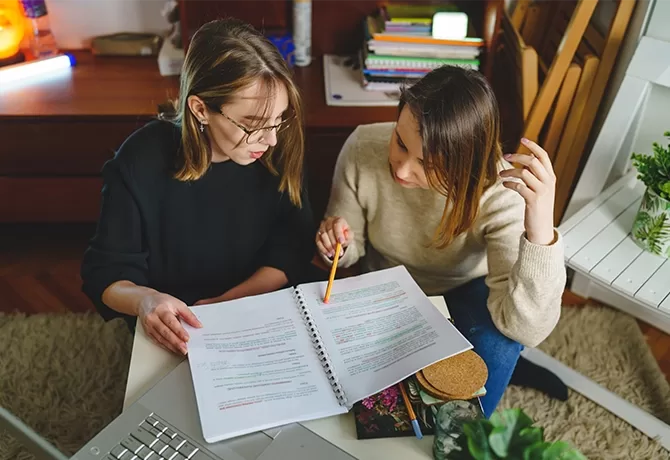 Two people are sitting at a table, focused on studying. They are looking at a notebook filled with highlighted text. One person is pointing at the notes with a pencil, and the other is listening attentively. Books and stationery are scattered around.