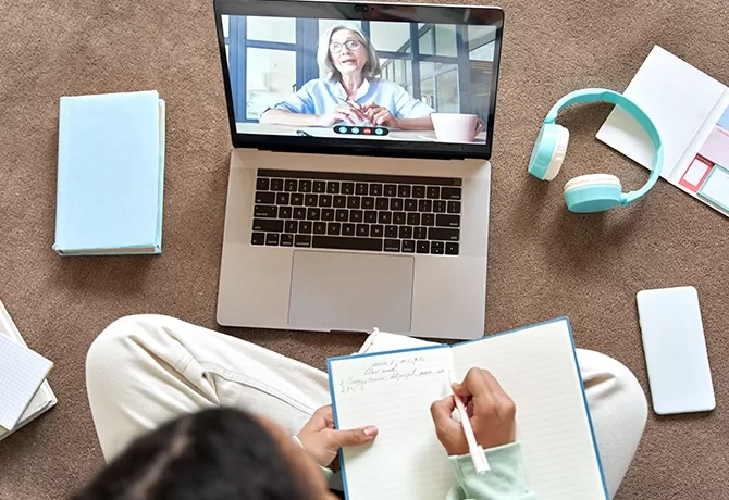 A person is sitting on the floor, taking notes in a notebook while attending a video call on a laptop. Headphones, books, and a smartphone are nearby. The laptop screen shows an older person speaking.