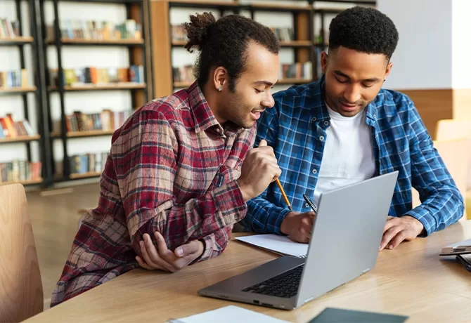 Two people sitting at a library table, working on a laptop. One is wearing a red plaid shirt, holding a pencil, and looking at the screen, while the other in a blue plaid shirt writes in a notebook. Shelves of books are visible in the background.