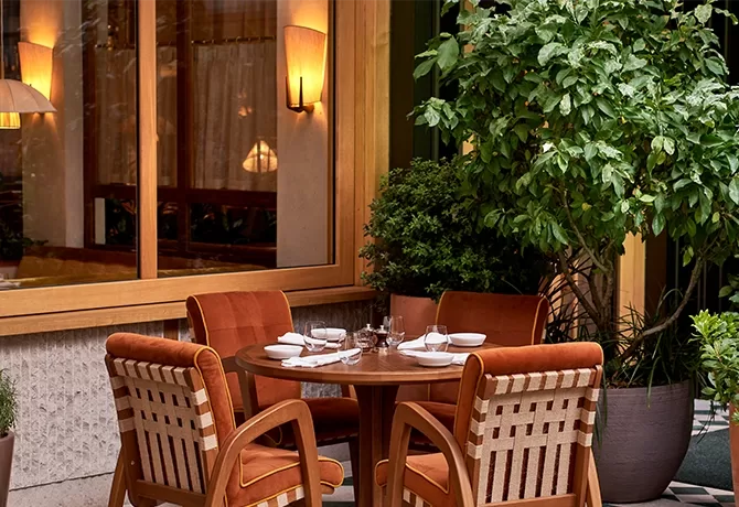 A cosy outdoor dining area features a round wooden table set for four, with burnt orange cushioned chairs. The setting includes white tableware and is surrounded by green plants, near a window with warm lighting at Dovetale, London.