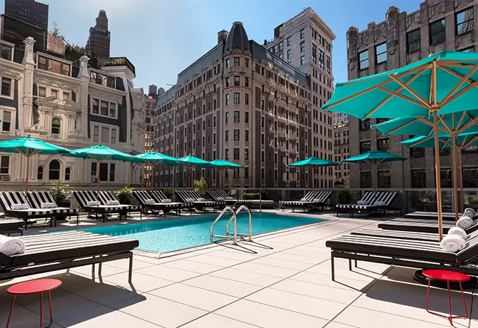Glimmering blue pool surrounded by New York buildings and turquoise parasols over sun loungers,