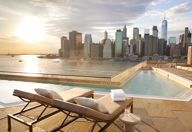 2 lounge chairs set out beside a pool overlooking the New York City skyline.