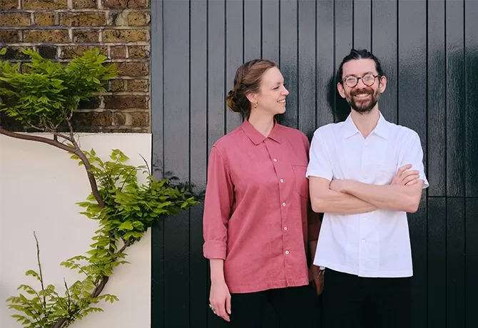 A woman in a red shirt smiles and looks at a man in a white shirt who stands with arms crossed. They are in front of a dark vertical wooden panel, with a green leafy plant climbing the wall beside them - the people are Desiree and John Chantarasak, chef's of the new restaurant AngloThai in London.