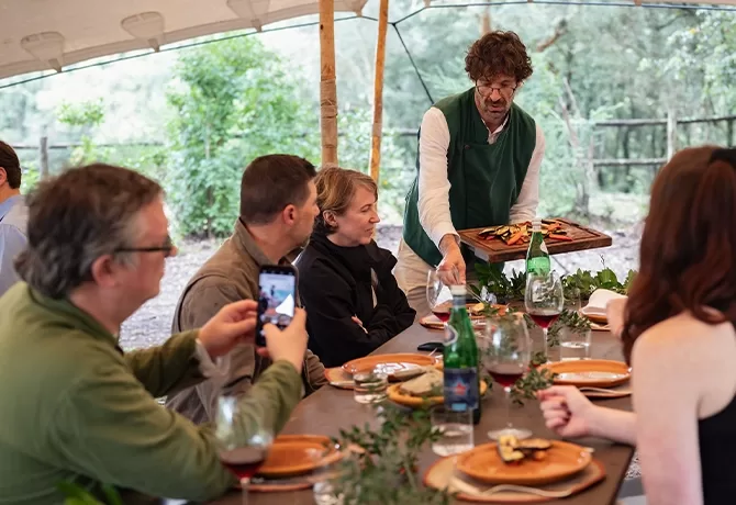 A group of people sit around a wooden table under a canopy, enjoying a meal outdoors. A server presents a platter of food. Wine glasses, dishes, and foliage decorate the table. One person is taking a photo with a smartphone.