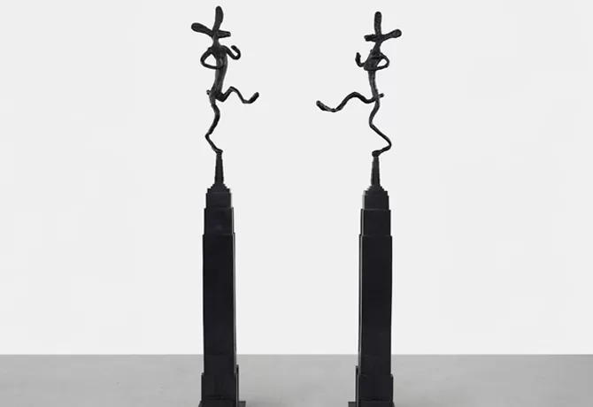 Two tall, slender black sculptures stand side by side, each topped with a whimsical, abstract figure in a dynamic pose. The design is minimalistic and stands on a flat surface with a plain background - Empire State with Bowler, Mirrored, 1997 by Barry Flanagan.
