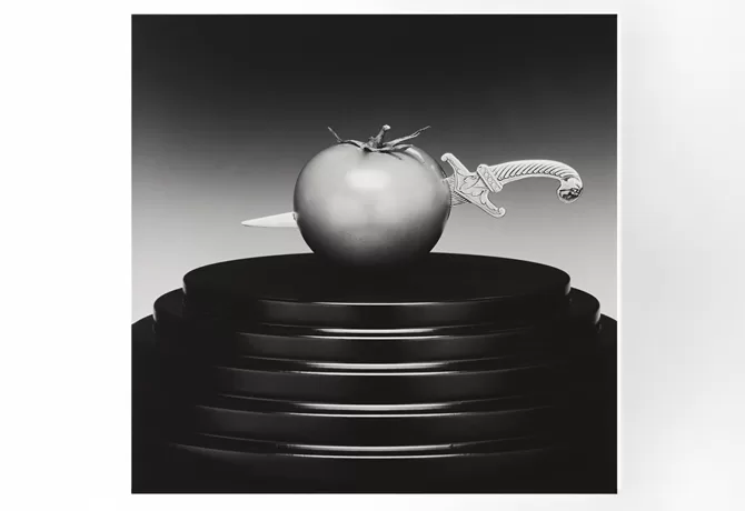 A black and white photograph of a tomato skewered by a decorative knife, displayed on a multi-tiered pedestal. The image contrasts the smooth tomato and intricate knife design against the stark background - Tomato/Knife, 1989 by Robert Mapplethorpe.