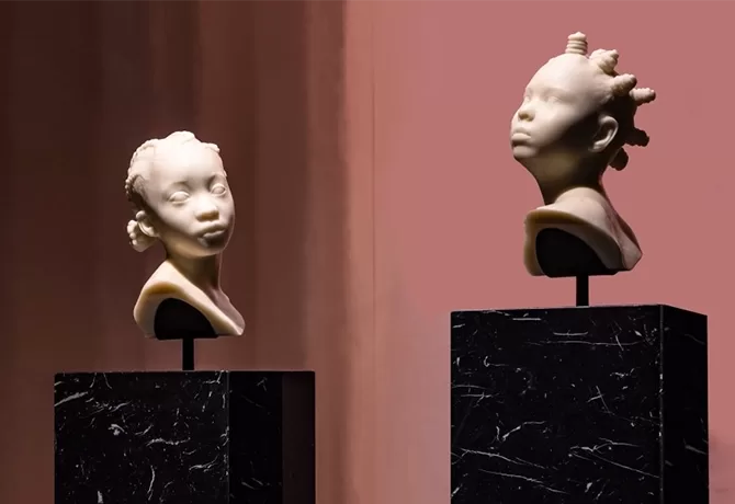 Two sculpted busts on black marble pedestals, depicting stylized portraits of children with unique hairstyles. The background features a warm, muted gradient - Ebony In Ivory III, 2023 and Ebony In Ivory II, 2023 by Esmaa Mohamoud.