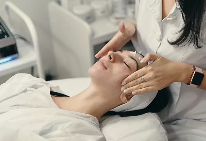 A person receiving a facial treatment in a spa setting. They are lying down with eyes closed, while a professional in a white coat gently massages their temples. Creams and skincare products are visible in the background.