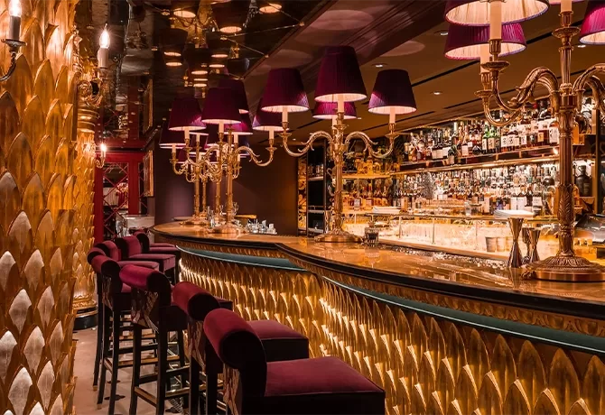 Luxurious bar interior with golden decor, ornate lighting, purple velvet bar stools, and a well-stocked mirrored bar counter, creating an elegant and inviting atmosphere.