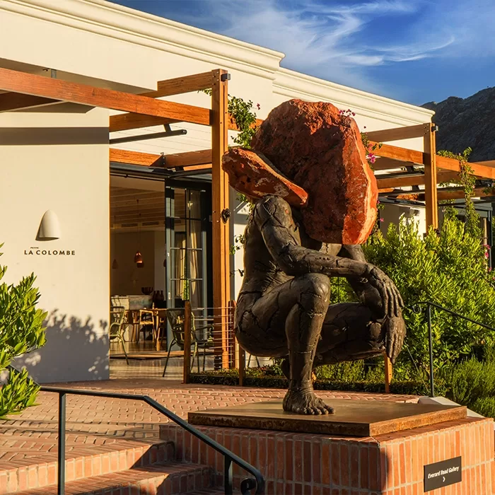 A bronze sculpture of a crouching human figure with a large, reddish stone obscuring its head is displayed on a brick platform outside La Colombe restaurant, surrounded by greenery and modern architecture.