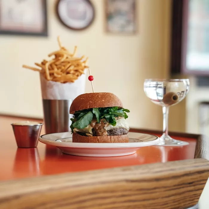 A cheeseburger with lettuce sits on a plate next to a metal cup of French fries and a martini with an olive, all arranged on an orange tabletop in a cozy restaurant setting.