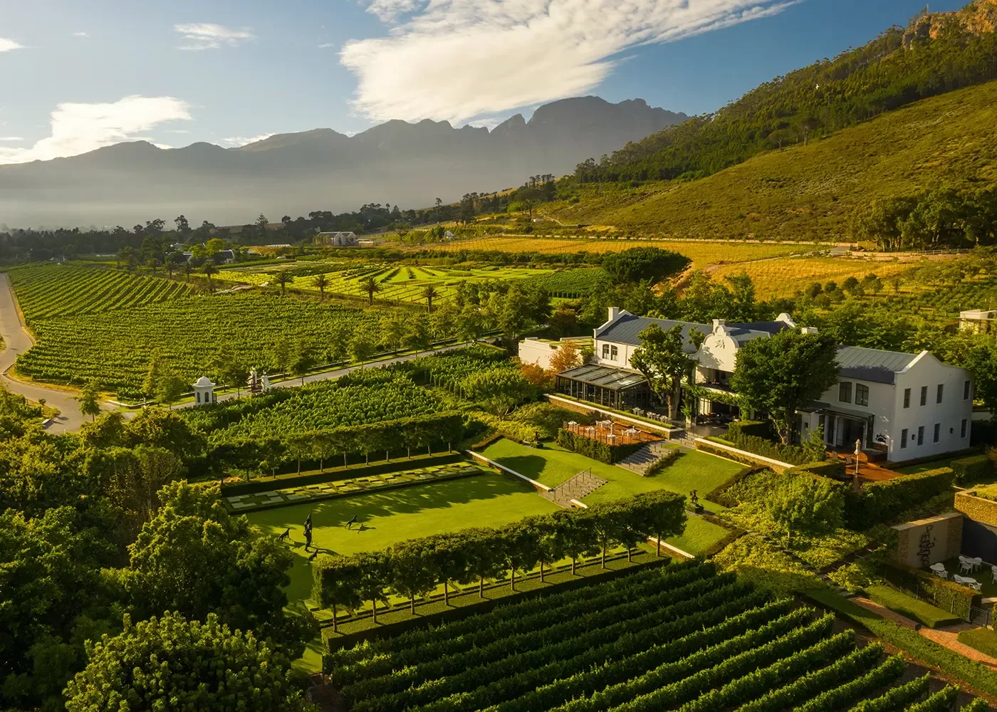 Aerial view of a lush vineyard estate with a white house, manicured gardens, and rows of grapevines, surrounded by green hills and distant mountains under a partly cloudy sky.
